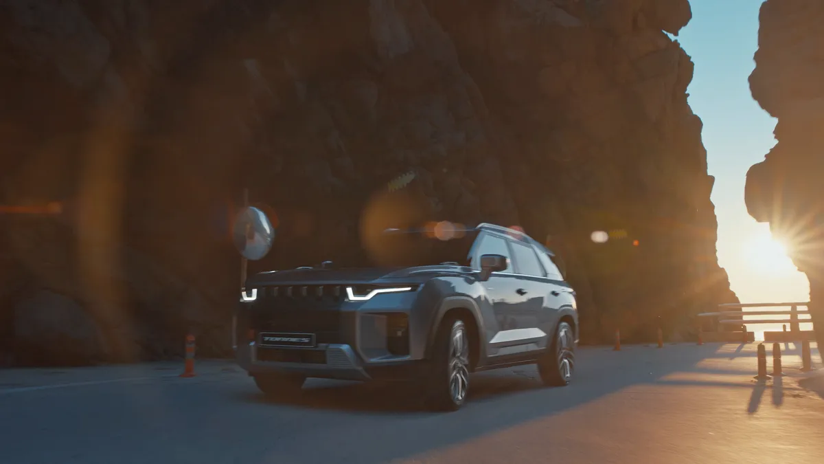 A grey KGM Torres driving out of a cliff-shaded coast road into the low evening sun